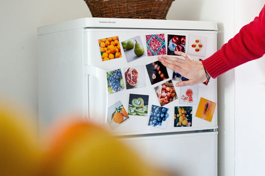 Hand placing fruit photo magnets on a compact white EnergySaver fridge in a Canadian kitchen, showcasing vibrant produce and small appliance style.